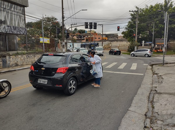 #PraCegoVer: Fotografia de uma profissional da saúde distribuindo máscaras no carro, ela esta de branco realizando às distribuições.